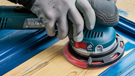 한 손이 나무 위에 전기 샌더를 들고 있으며, 파란 작업대에 둘러싸여 있습니다.