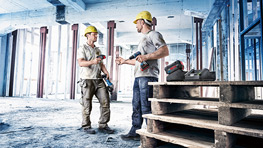 헬멧을 쓴 두 건설 노동자가 건설 현장에서 서 있습니다. 그들은 토론 중이며, 도구가 팔레트 위에 놓여 있습니다.