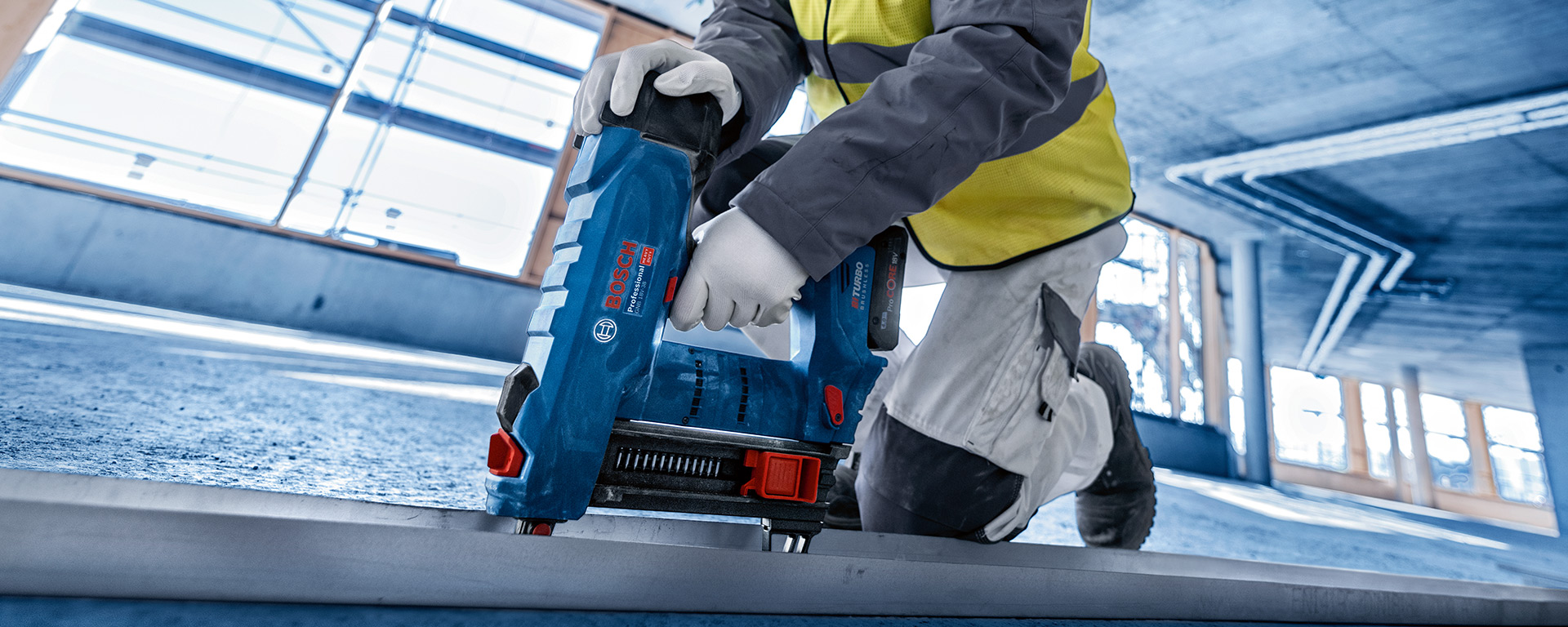 노란 조끼를 입은 작업자가 바닥에서 파란색 Bosch 못 박기를 사용하고 있습니다.
