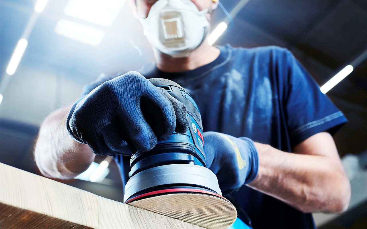 파란 장갑을 낀 장인이 Bosch 장비로 나무를 연마하고 있습니다.