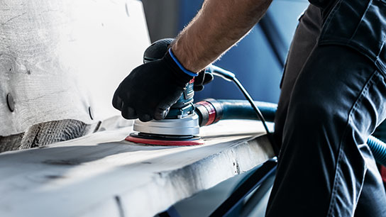 장갑을 낀 장인이 나무에 보쉬 연마기를 사용하고 있습니다.