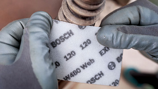 회색 장갑을 낀 손이 나무 표면 앞에서 Bosch 사포를 들고 있습니다.