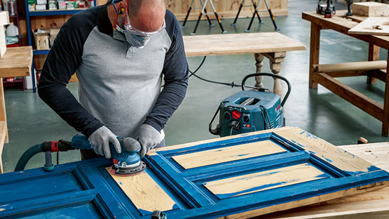 한 남자가 작업장에서 Bosch 샌더로 파란색 문을 샌딩하고 있습니다.