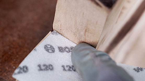 회색 장갑을 낀 손이 사포로 나무 모서리를 갈고 있습니다.