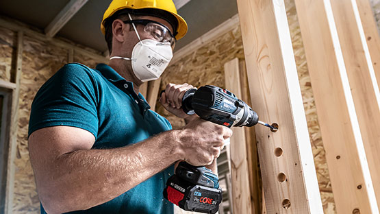 헬멧과 마스크를 쓴 남자가 Bosch Professional 무선 드릴로 나무를 뚫고 있습니다.