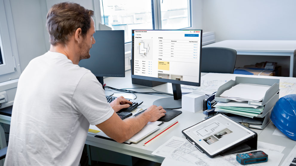 한 남자가 건축 계획이 있는 밝은 사무실에서 컴퓨터로 작업하고 있습니다.