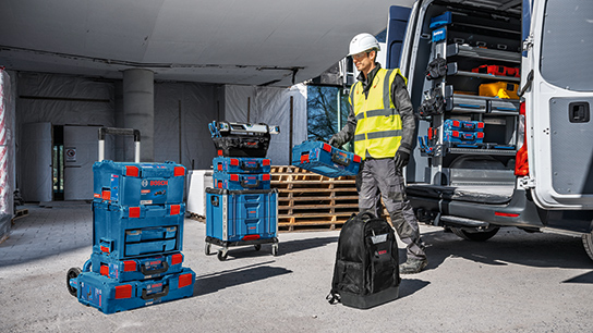 안전 조끼와 헬멧을 쓴 작업자가 밴 옆에 서 있고, 파란색 Bosch Professional 공구 상자들로 둘러싸여 있습니다.