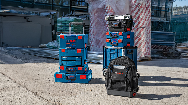 여러 개의 쌓을 수 있는 파란색 Bosch Professional 공구 상자가 검은색 배낭 옆에 건설 현장 바닥에 놓여 있습니다.
