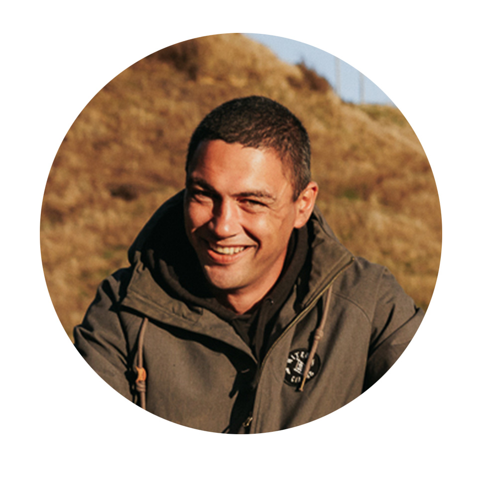 회색 파카를 입은 웃고 있는 남자가 풀밭 앞에 앉아 있습니다.