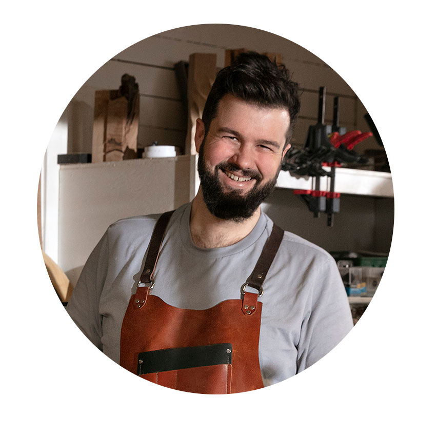 아담 데트레, 조각가, 아담 데트레 디자인 가죽 앞치마를 입은 웃고 있는 남자가 작업실에 서 있습니다.