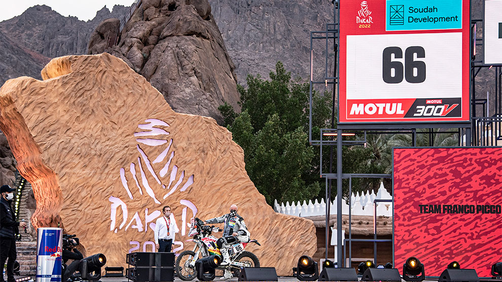 오토바이 운전자가 사막의 큰 다카르 표지판 앞에 서 있습니다.