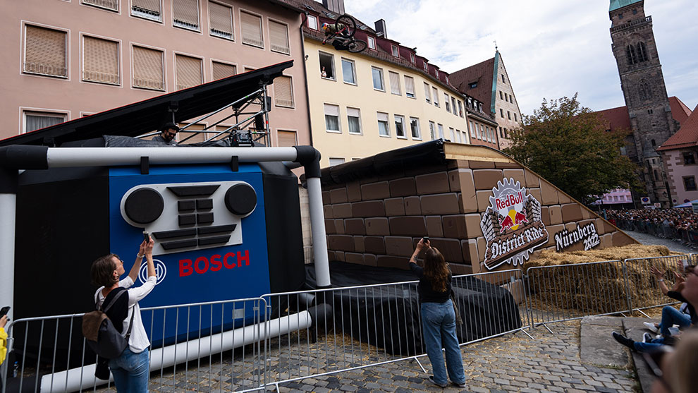 거대한 보쉬 업 박스에서 도약한 라이더가 공중에서 기술을 선보이고 있습니다.  관중, 스턴트 자전거 및 역사적인 건물이 배경에 있는 Bosch 광고 설치.