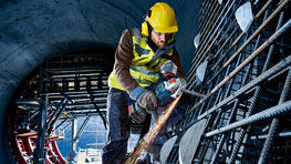 노란 헬멧과 경고 조끼를 입은 작업자가 금속 막대를 자르고 있습니다.